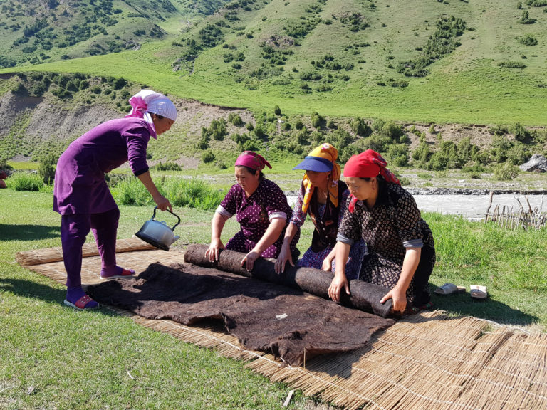 Four Kyrgyz women working together on felt-making in a mountainous grassland, a craft deeply rooted in craft markets in Central Asia. One pours water from a kettle while the others roll and press dark felt on reed mats, with lush green hills in the background.