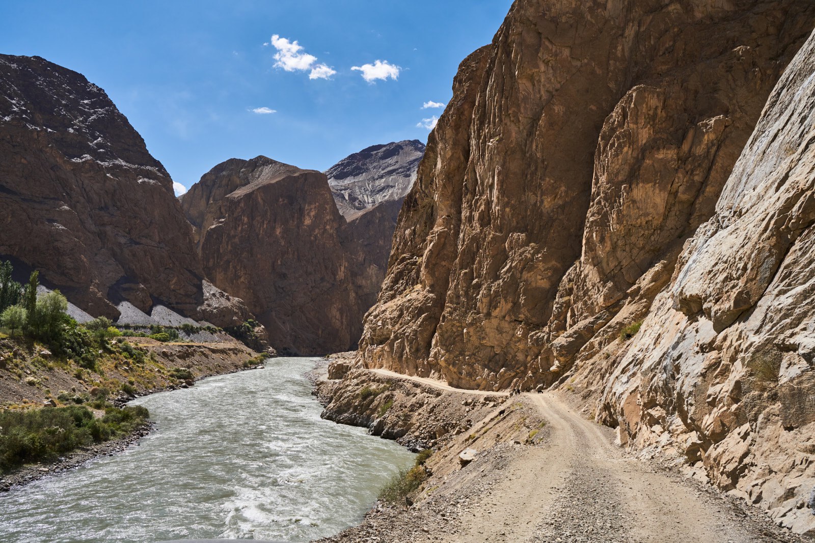 A dramatic mountain gorge in the Pamir Mountains, with steep rocky cliffs rising above a narrow dirt road that hugs the cliff face