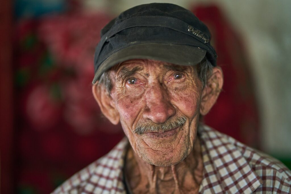 Close-up portrait of an elderly man with weathered, wrinkled skin wearing a dark cap and a red and white checkered shirt