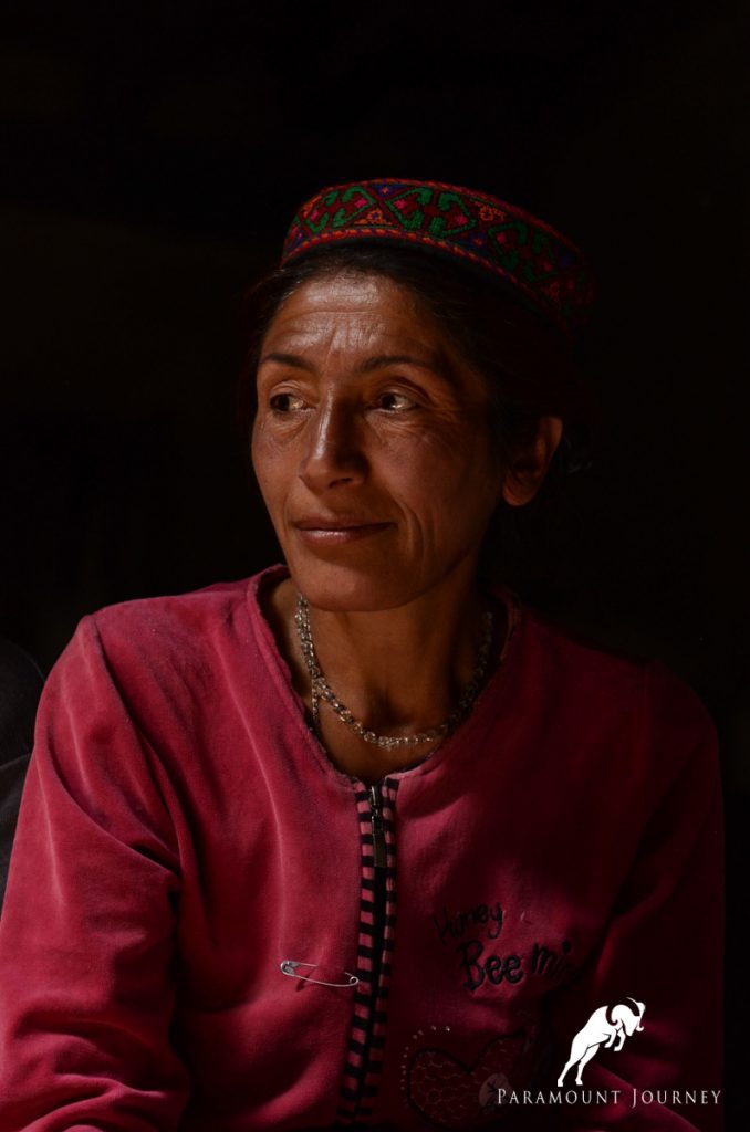 A middle-aged woman wearing a traditional embroidered Pamiri cap and a red sweatshirt smiles gently while looking to the side