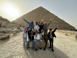 Paramount Journey team posing cheerfully in front of a large pyramid on a sunny day. The group, consisting of men wearing casual attire, some with traditional scarves, is raising their hands in celebration. They are seated and standing on a rocky surface near the iconic ancient structure