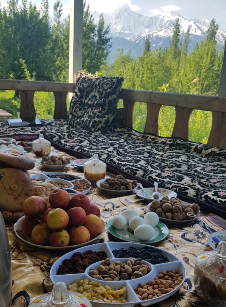 Traditional Tajik dastarkhon with dried fruits, nuts, fresh apples and eggs set against snow-capped Pamir mountains, showcasing authentic mountain hospitality