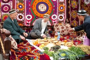 Traditional Tajik elders sharing a meal on dastarkhon with fresh bread and fruits, seated against colorful suzani tapestry displaying authentic Tajik hospitality