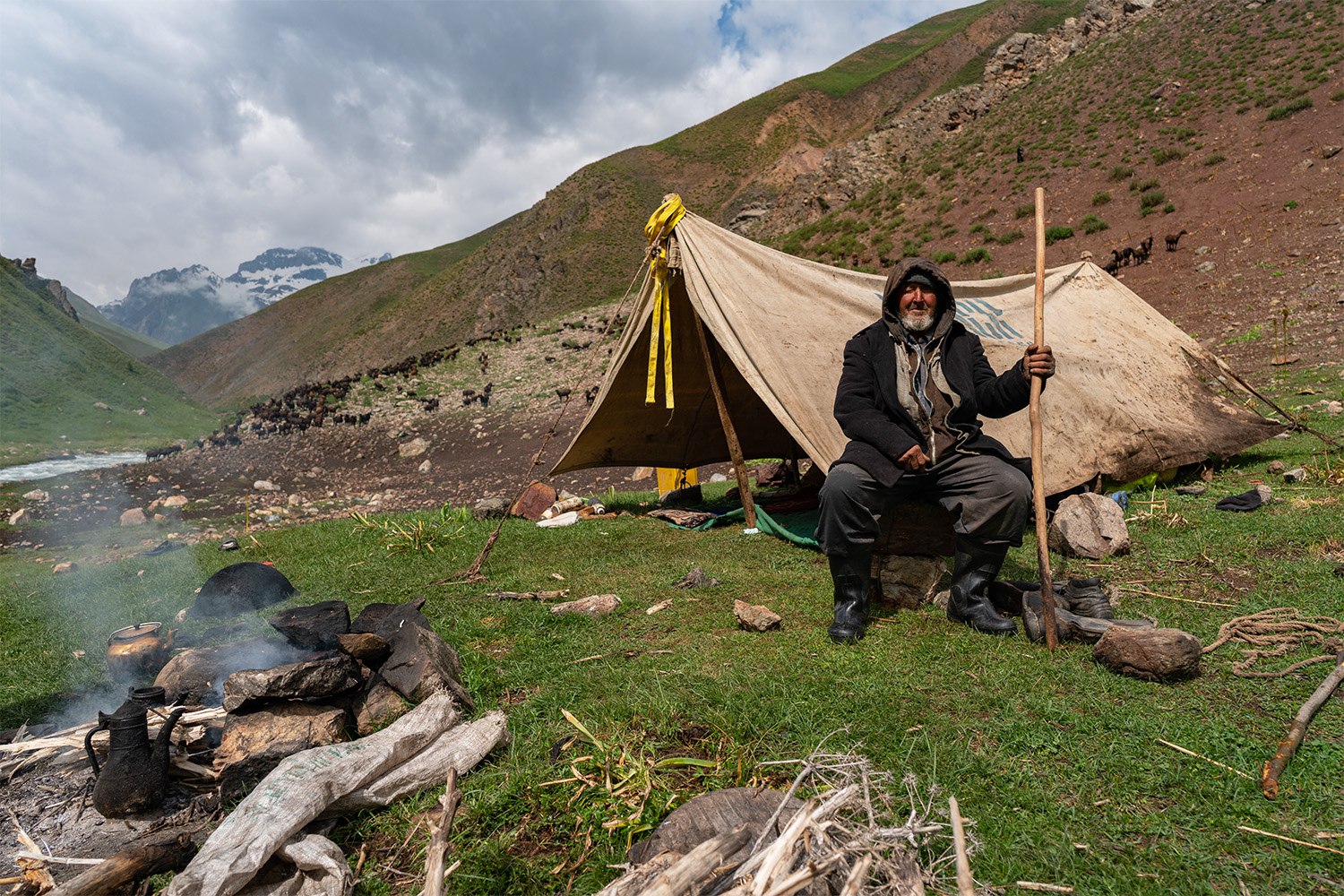 Shepherd sits outside his tent camp with smoking campfire in foreground and snow-capped mountains in background