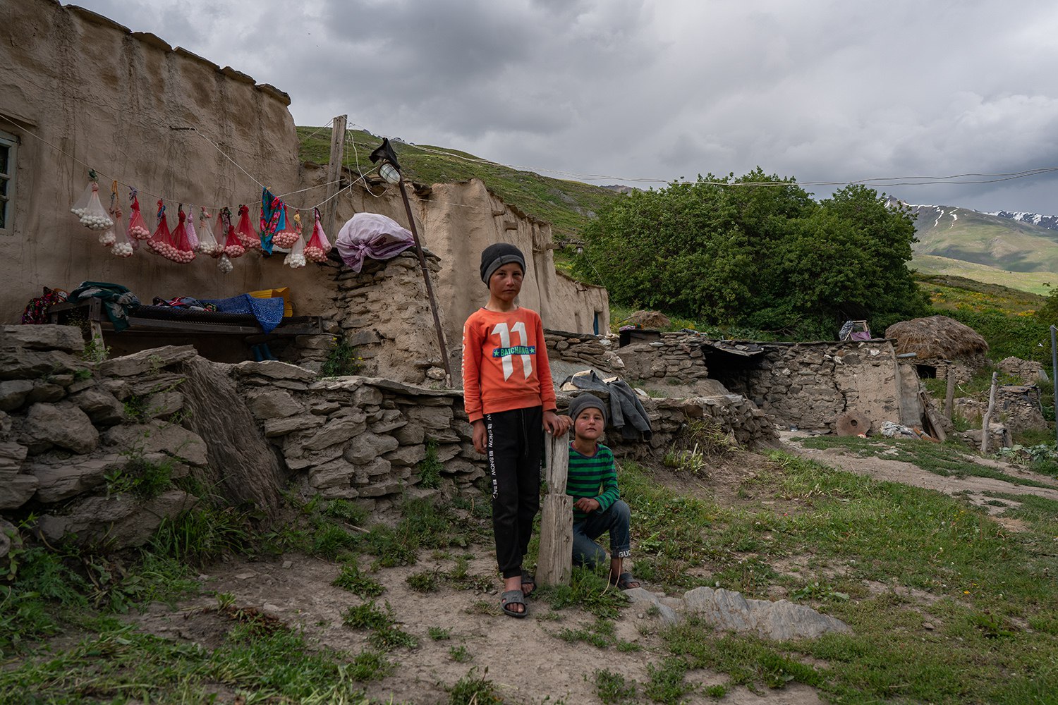 Two children standing outside a traditional stone and mud house with a backdrop of green hills, trees, and cloudy skies in Yagnob, Sogd