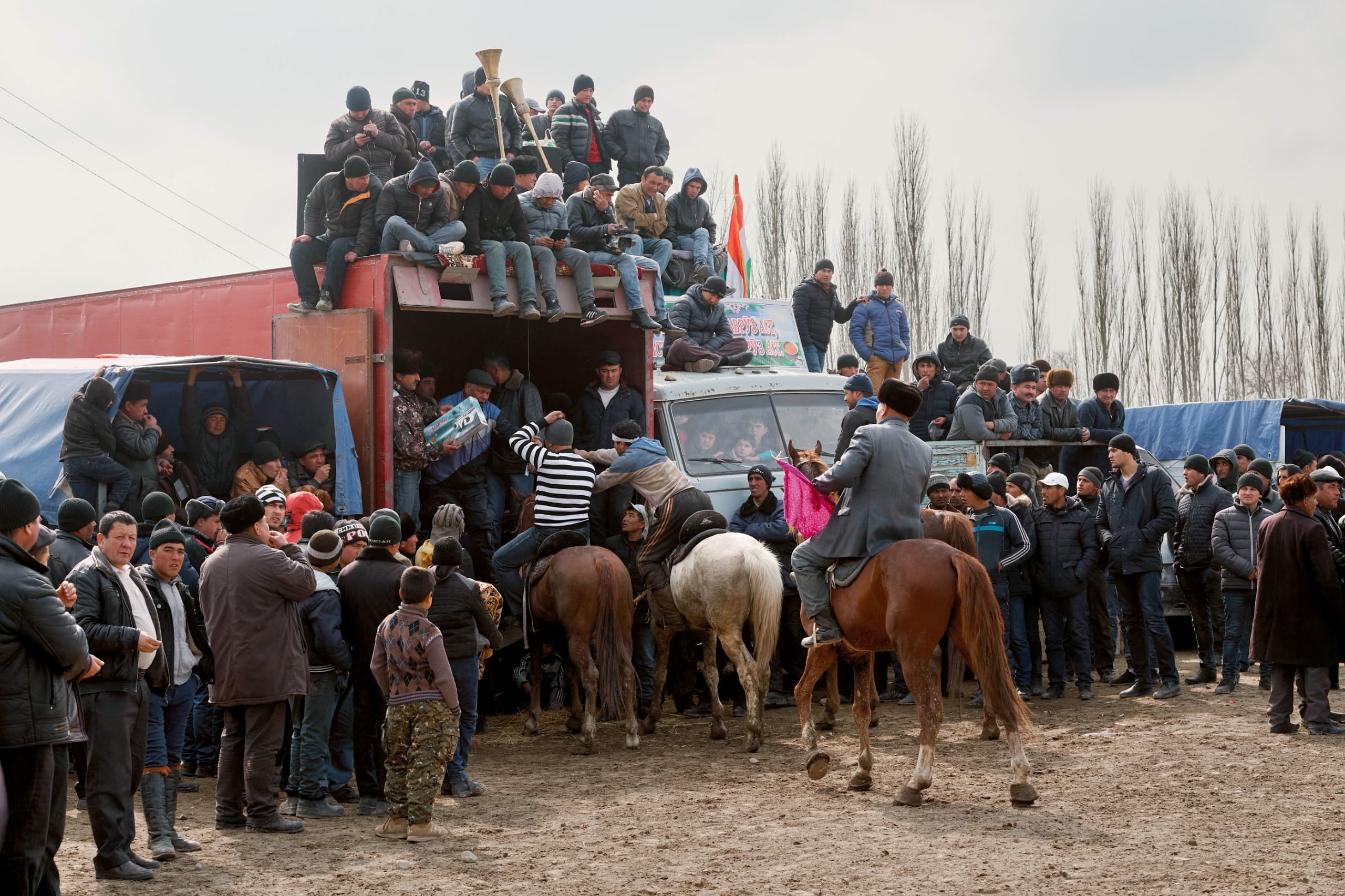 A lively prize ceremony following a Buzkashi match in Tajikistan, where a crowd gathers around trucks serving as makeshift stages. Men on horseback approach the distribution point to receive awards, while others climb onto or sit atop the trucks. Some spectators hold musical instruments, and one person hands over a box of detergent as a prize.