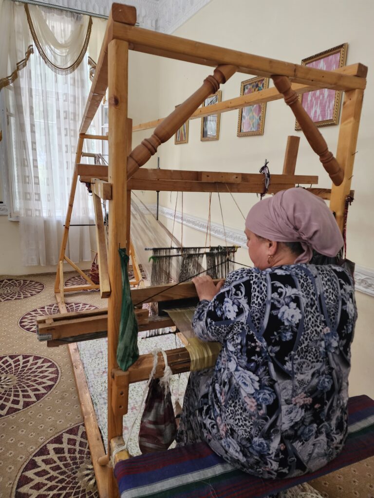 A woman wearing a lavender headscarf and floral patterned clothing works at a traditional wooden handloom in a home setting in Khujand. She is seated on the floor weaving atlas fabric, with framed samples of ikat textiles displayed on the wall behind her. The wooden handloom features a traditional design with multiple foot pedals.