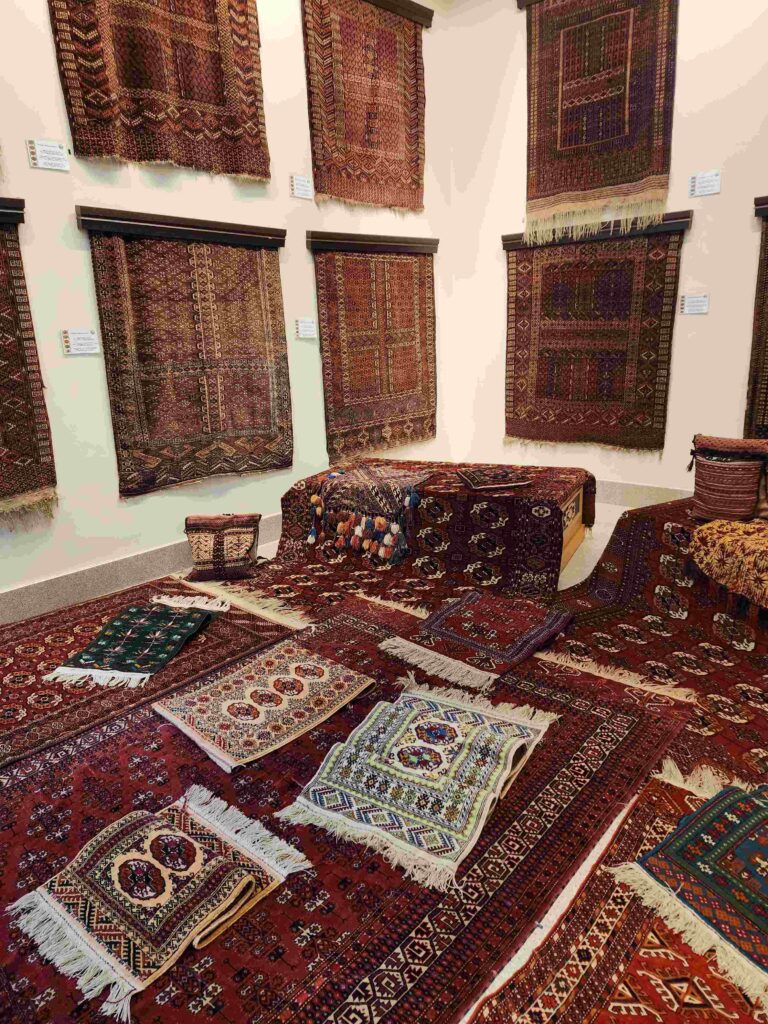 A display of traditional Turkmen carpets and rugs in a museum or gallery setting. The carpets, predominantly red with intricate geometric patterns, are hung on the walls, while smaller rugs and textiles are arranged on the floor and furniture.