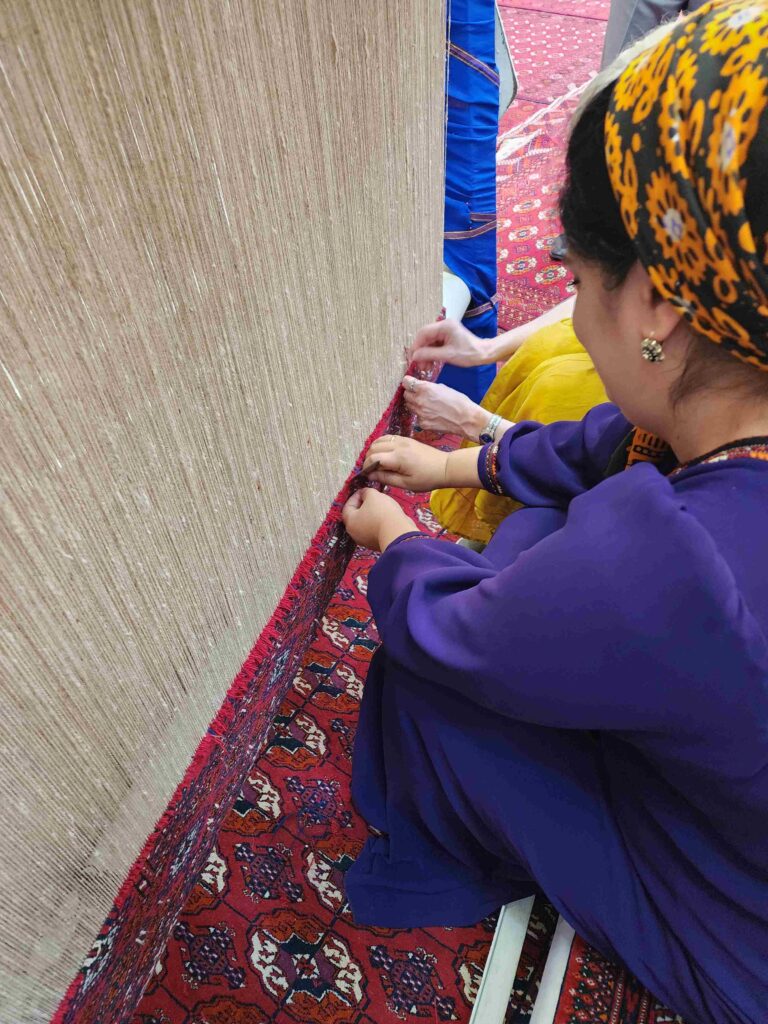 Two women weaving a traditional Turkmen carpet on a loom. One woman, dressed in a purple outfit with a yellow-patterned headscarf, is tying knots on the vertical threads of the loom, while another person in yellow assists. The background features a red-patterned carpeted floor, characteristic of Turkmen designs.