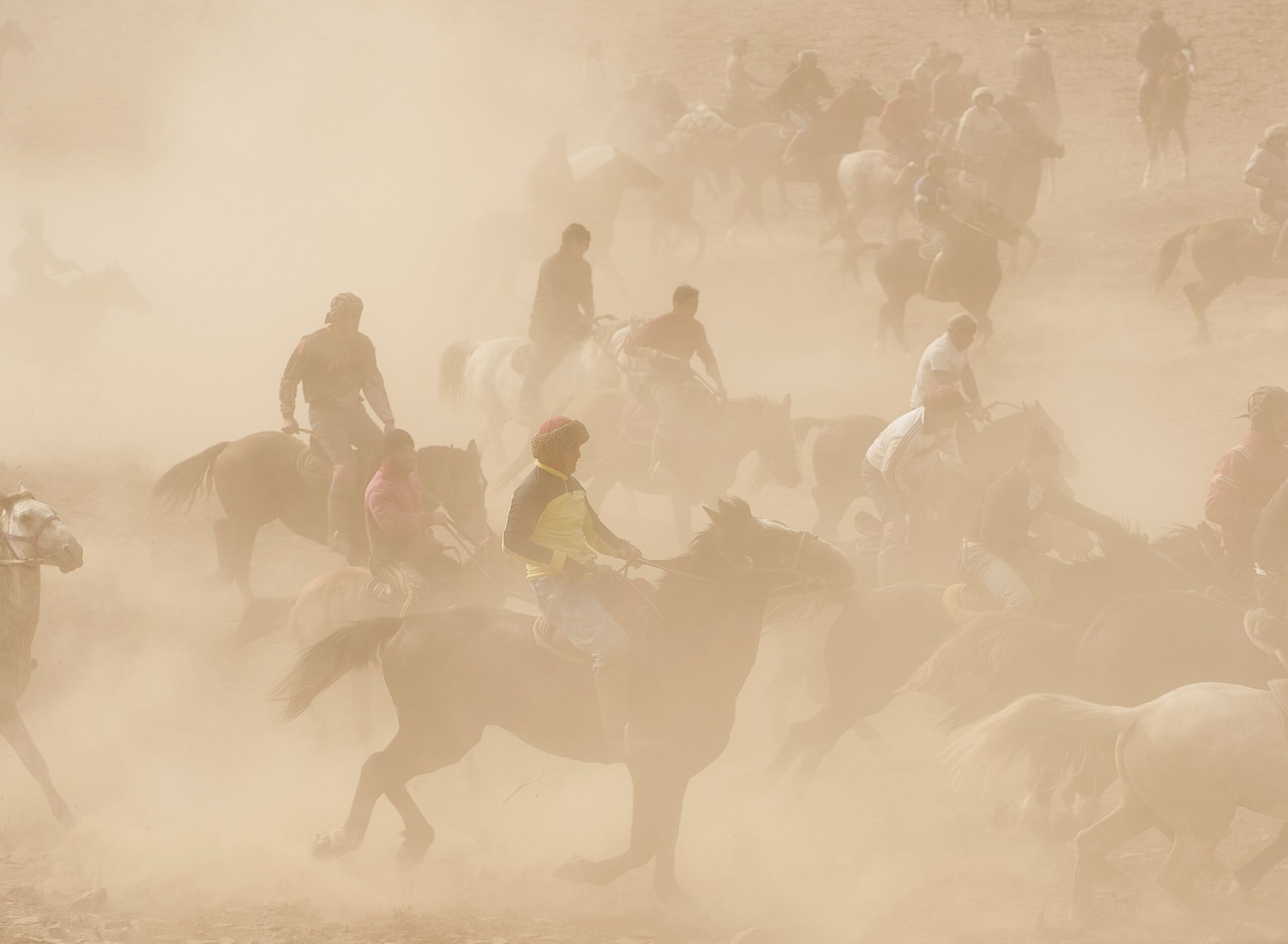 Buzkashi players on horseback competing in a dust cloud during traditional equestrian sport, with riders wearing colorful traditional clothing including yellow and white garments