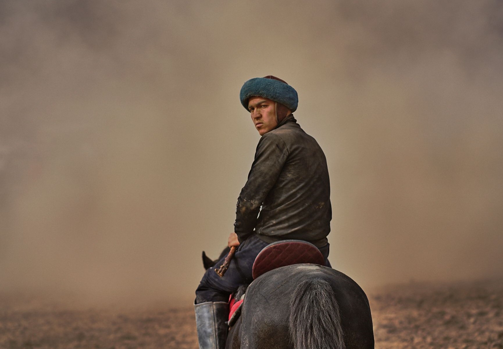 Buzkashi rider wearing traditional blue hat on dark horse with red saddle, portrait shot during equestrian competition with dust clouds in atmospheric background